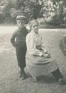 Young blond boy with newsboy cap, approximately age 7, stands next to his seated sister, who is wearing a blouse and skirt, for a posed photograph taken outdoors
