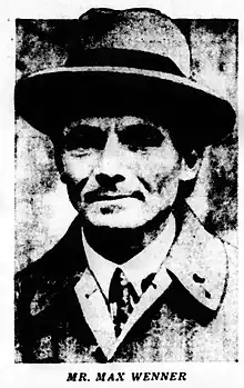 Grainy black-and-white headshot of a clean-shaven man wearing a suit and tie, overcoat and hat