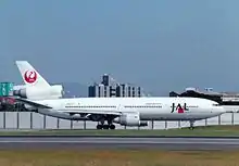 A McDonnell Douglas DC-10 aircraft taxiing on the tarmac, with a yellowish grass strip in the foreground and buildings and fence in the background