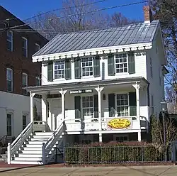 A two-story white wooden house with decorative touches, a pointed metal roof and green shutters. A sign in the center of the porch reads "Irvington History Center" while another at the right on the story below says "Frocks, Frills and Furbelous 1760-2009"