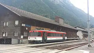 Red-and-white train at side platform in front of large three-story wooden building with gabled roof