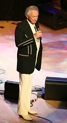 A grey-haired man in a black jacket and white pants, singing into a microphone