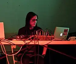 Merzbow, a Japanese man with long black hair wearing a dark t-shirt, dark pants, and sunglasses, sits behind a table looking at a soundboard and MacBook.