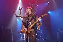 Long-haired man in beige shirt with wide sleeves and a brown leather vest and pants, plays the electric guitar on stage and sings. There is a large pair of felt antlers pinned to the microphone.