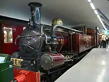 A steam tank locomotive is shown indoors, funnel towards the viewer, in purple livery. A large pipe connects the pistons at the front with the side tank