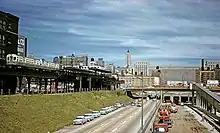 A view looking northeast into downtown Chicago. An elevated railway station, somewhat dilapidated with brown peaked canopies on its platforms, is depicted on the center left with a train of two cream-and-green cars approaching its southern, eastbound platform. Just right of center, below the elevated structure, is an expressway with train tracks in the middle of it and a train station's newly constructed ramp descending into it; only the northern, westbound, track is depicted; the rest is cut off by the edge of the image.