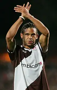 African man in a diagonally striped brown-and-white jersey with a sponsor logo across the chest. His hair is cornrowed, and his arms are crossed above his head.