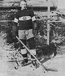 A man stands outside in skates, wearing a sweater with a team logo on it, and wears gloves and holds an ice hockey stick.