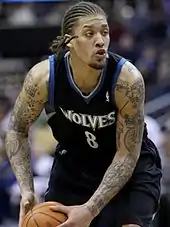 Michael Beasley with cornrows, wearing a black Timberwolves uniform with blue trim and griping a basketball with both hands in the lower left