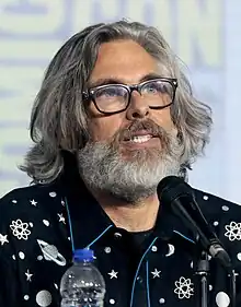 Michael Chabon is pictured wearing glasses, shoulder-length hair and a beard. Speaking at a microphone, his collared shirt is decorated with celestial bodies.