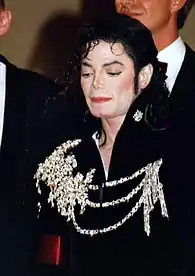 Jackson is wearing a black velvet jacket with a crystal rhinestone metal appliqué in relief. It covers his right shoulder. Three lines of rhinestones cross his chest and gathering on the left side. The neck is unbuttoned. A rhine stone crown adorns the turned up collar. He's looking down. His skin is light and he's wearing makeup.