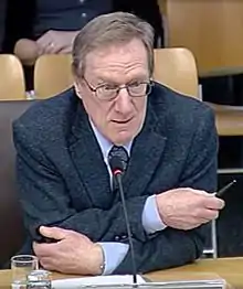 close-up of Michael Keating wearing a light blue shirt and dark blazer, leaning towards a microphone on a desk, appearing to speak