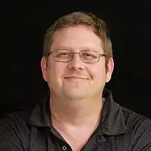 a white man with short brown hair wearing a black polo and rectangular glasses smirks