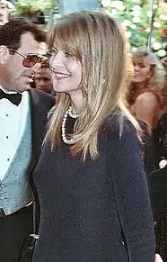 Young Caucasian woman with long blonde hair, wearing a black dress and pearl necklace. She smiles politely.