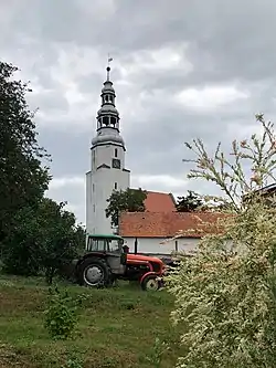 The tall white tower of the church in Mierczyce village having a clock face and standing near the red roof of the church as seen across a nearby yard where a tractor being parked in.