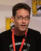 A man with dark brown hair and glasses leaning slightly forward, and speaking into a microphone.