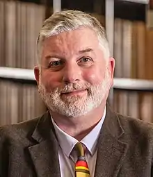 Portrait photograph of a bearded man wearing a brown corduroy sport jacket, blue shirt, and brown tie with red, yellow, and blue stripes. The photo is framed from the upper chest to just before the edge of either shoulder with blurred files in the background, creating a pleasing boke effect.