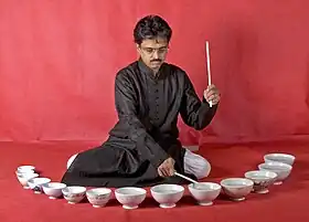 Milind Tulankar sitting on the ground with water bowls laid out in front of him as he plays music on them