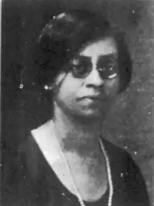 A Black woman with jaw-length dark hair and glasses, wearing a dark scoop-neck dress and a strand of pearls