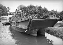 Four shirtless men in a large barge moored at a river bank. The barge has a ramp at the front of a catamaran hull.