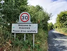 road sign (tempo limit 30): Welcome to MINIONS, Please drive carefully; on each side of the lettering is a very faded sticker of a Despicable Me "minion"