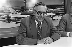  Max van der Stoel sits smiling at a table in an airport. Planes can be seen behind him.