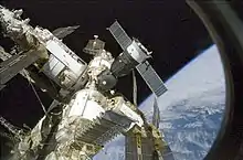 A cluster of modules, covered in white insulation and projecting feathery solar arrays, with a small spacecraft covered in brown insulation docked at their centre. The image is seen through a window, with the blackness of space and the Earth forming the backdrop.