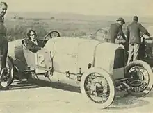 Black and white photo of a white woman in an old race car. Several men stand around.
