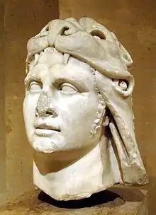 Slightly damaged stone sculpture of a man's head. He wears an animal pelt over his hair.