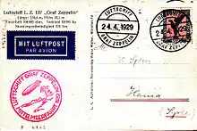 The reverse of a postcard. At the top left is the name of the airship and some of its performance statistics. Below is a dark blue rectangular sticker saying "Mit Luftpost Par Avion". Below that is a circular red cachet saying "Luftschiff Graf Zeppelin Mittelmeerfahrt 1929", around a stylised airship flying from right to left above a desert landscape with a palm tree. At the top right the stamp has been cancelled by a circular postmark saying "Luftschiff Graf Zeppelin 24.4.1929". The postmark is repeated to the left of the stamp. The consignee's address is in Hama, Syria, and is hand-written.