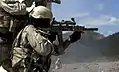 US Army SF takes aim with his desert camouflage-painted SPR. An Insight Technologies AN/PEQ-2A Target Pointer/Illuminator Aiming Light (TIPAL) is mounted on the right of the rifle's handguards. A standard M4 telescoping stock is mounted.