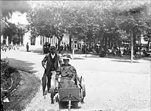 A scene from 1899 Luchon, with a woman in a wheelchair accompanied by a male attendant.
