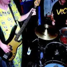 A low quality photo of a person in a SpongeBob onesie playing bass and someone else playing a drum set