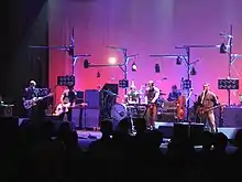 A group of six musicians performing on a stage, with an audience in the foreground and a gradient of pink and purple on a wall in the background.