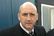 A middle-aged, balding man in a dark suit looks at the camera with a neutral expression on his face.