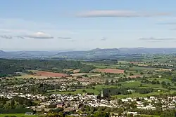 The image is taken from a height, and looks down on the town of Monmouth in the foreground. The buildings are mostly white, with slate roofs, and the only tall structure is the church spire. Beyond are some suburbs, giving way to rolling green fields and woodland. In the distance the Black Mountains are visible. Besides Sugar Loaf, which is sharply pointed, they are low and broad.