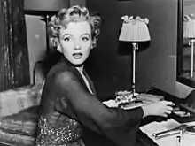  Monroe, wearing a transparent lace robe and diamond earrings, sitting at a dressing table and looking off-camera with a shocked expression