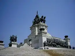 Photograph of a white stone steps leading up to a large, altar-like monument in white marble with bronze sculptural decorations that include bronze braziers at the corners, a bronze frieze in high relief at the base and bronze figures surrounding a chariot on a high, white marble plinth in the center