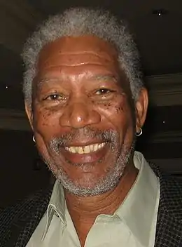 An African American man with a beard and short hair, both a mix of white and grey, and wearing an earring in each ear: He is smiling towards the camera.