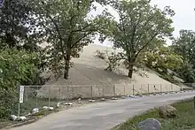 A metal fence is shown in front of a massive dune which is overtaking the road.