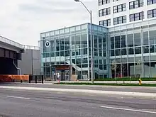 Photo of a glass station building with an entryway at ground level. An elevator and staircase are visible inside.