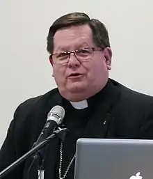 A bespectacled man wearing a black cassock and Roman collar