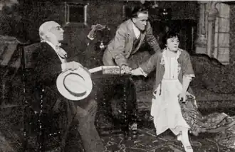 stage scene: seated left, an old man with receding white hair, gesticulating; a younger man, standing, and a woman, seated looking horrified