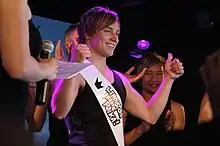 Man wearing a black T-shirt and white sash smiling and giving a thumbs-up