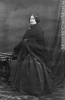 A black and white photograph of a woman with dark hair in a long, dark dress with a thick shawl.