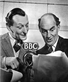 two middle aged, clean-shaven white men, one with full head of dark hair and the other bald; they are in front of a microphone in a recording studio, reading a script