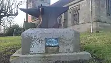 An anvil on a pedestal, with a stone church in the background. The pedestal has a plaque reading "This anvil came from the smithy when it was demolished and is believed to have been used by William Skelhorn in 1459"