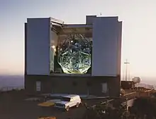 A rectangular building about the size of a small apartment block stands on a mountaintop, with a vehicle parked in front of it. The building has a wide opening in its front and top, exposing an internal framework structure that holds and somewhat obscures from view a hexagonal arrangement of six circular primary mirrors. In the background, the sky is hazy, hiding the horizon and most of the terrain below the mountain.