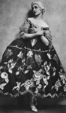 Muriel Stuart, an English ballerina in 1921, wearing a dark and voluminous costume with floral embroidery, and a powdered wig. She has her hands crossed at her chest. One foot is visible.