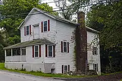 A white wooden house with clapboard siding and a front-gabled roof, seen at a three-quarter angle from across a road in front of it. Its windows are in an irregular pattern, with red wooden shutters and double-hung 12-over-12 sash windows. A porch comes off to its left to wrap around the far side of the house, and there is a door between the two windows on the second story facing the road. On the side a large brick chimney rises to the roof.